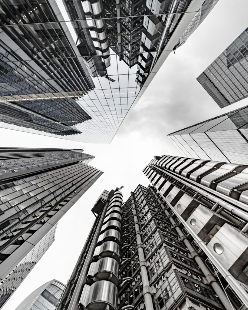 Esta imagen muestra una vista arquitectónica hacia arriba entre rascacielos modernos de acero y vidrio, transmitiendo sensaciones de poder corporativo, innovación y dinamismo urbano. Es ideal para ilustrar temas de crecimiento inmobiliario, inversiones o transformación digital en el sector inmobiliario.